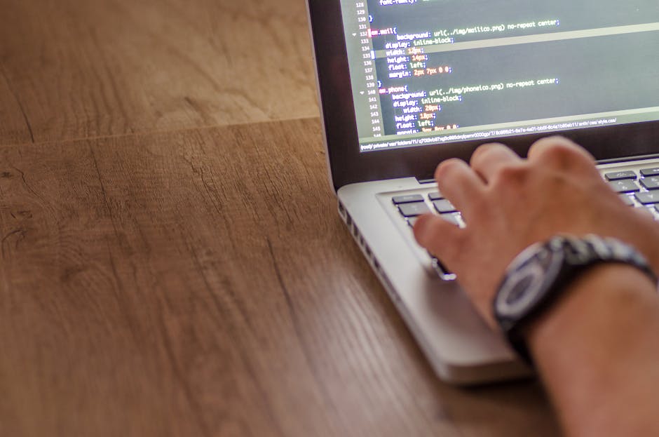 A person's hand, wearing a wristwatch, is seen typing on a laptop keyboard placed on a wooden desk. The laptop screen displays lines of code with colorful syntax highlighting. The desk has a smooth, n