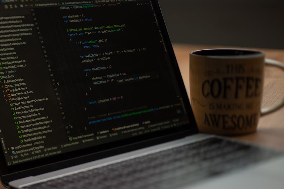 A close-up photograph of a laptop displaying lines of colorful code on its screen, positioned on a wooden desk. In the background, a ceramic mug with the phrase 'This coffee is making me awesome' in b