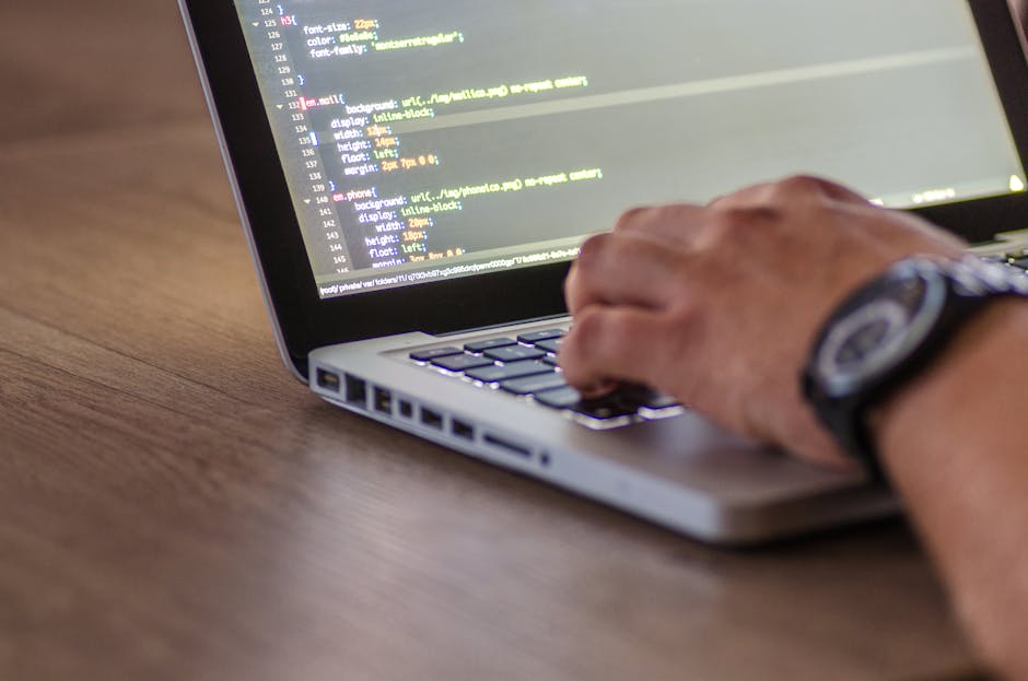 A close-up photograph of a laptop displaying lines of colorful code on its screen, positioned on a wooden desk. In the background, a ceramic mug with the phrase 'This coffee is making me awesome' in b