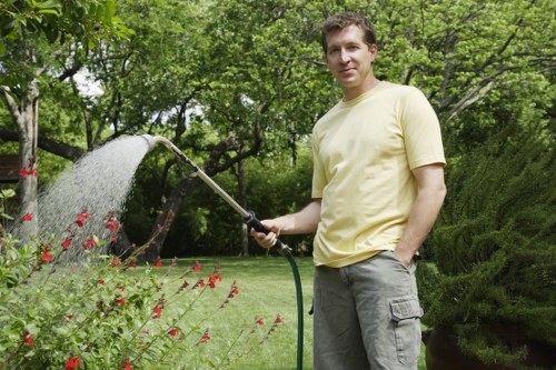 A man in a pale yellow t-shirt and grey cargo shorts stands in a well-maintained garden in Muswell Hill, London, holding a garden hose that waters a lush flower bed filled with red blooms. The garden features a neatly trimmed lawn with dense, green grass, bordered by a variety of shrubs and trees, including an evergreen hedge in the background. Sunlight filters through the leafy canopy, casting natural light over the scene, indicative of a pleasant weather day. The garden setting showcases typical outdoor landscaping elements such as flower beds, grass turf, and mature bushes, highlighting a cared-for, inviting outdoor space suitable for gardening services provided by Gardener Muswell Hill. The scene emphasizes the healthy, vibrant appearance of the garden, reflecting regular maintenance and landscaping work, with a focus on natural tones of green, red, and earthy soil. The garden appears spacious and well-structured, with visible plantings and edging that create a pleasing environment for outdoor enjoyment and gardening care in the Muswell Hill area near N10 postcode.